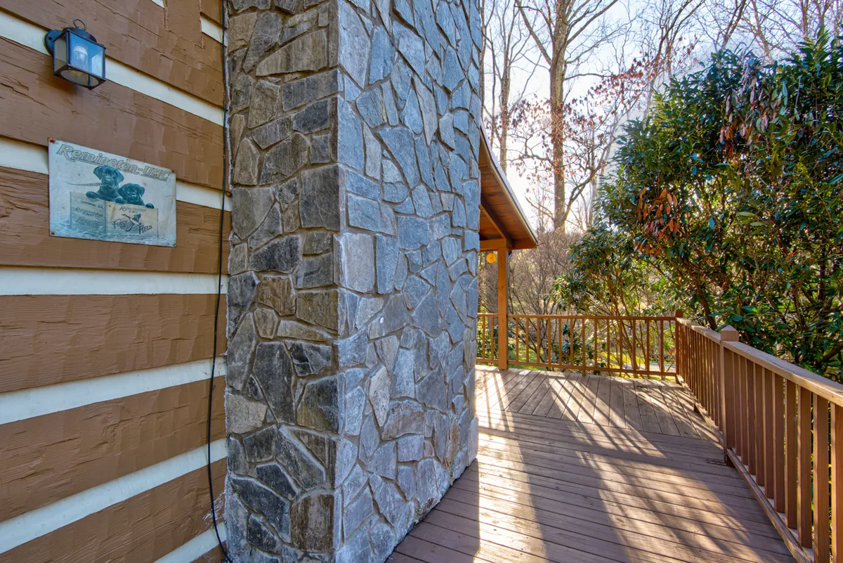 A warm exterior view with a stone wall, wooden deck, and cabin signage. The deck offers space for outdoor relaxation, gatherings, and enjoying sunset views, framed by the surrounding trees.