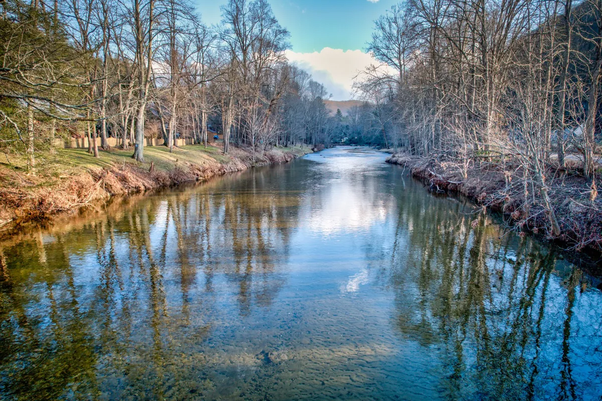 A serene riverside view featuring a calm river, reflections of trees on the water, and a tranquil sky. The peaceful setting is ideal for storytelling, picnics, and group bonding experiences by the water's edge.
