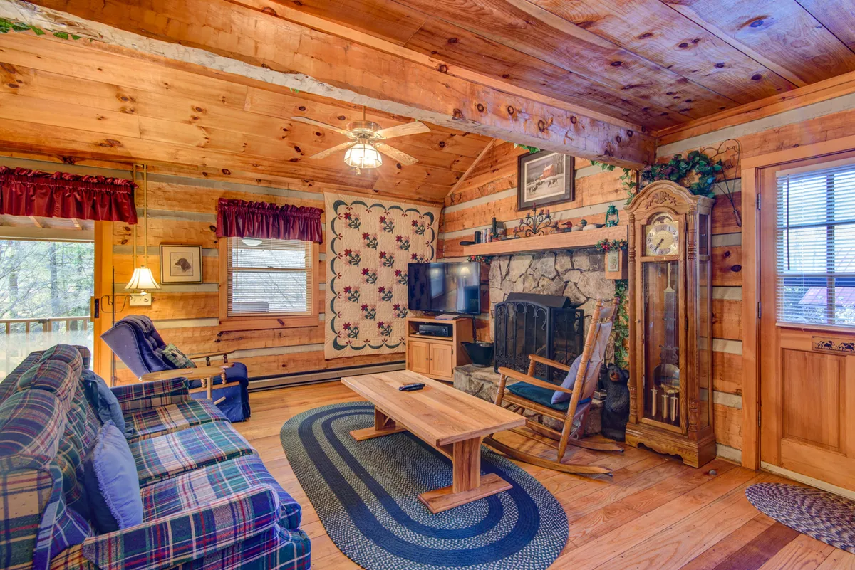 A comfortable family room centered around a stone fireplace, with a plush sofa, coffee table, wall quilt, television, and a classic grandfather clock. The decor feels inviting and homey, ideal for relaxing or playing games as a family.