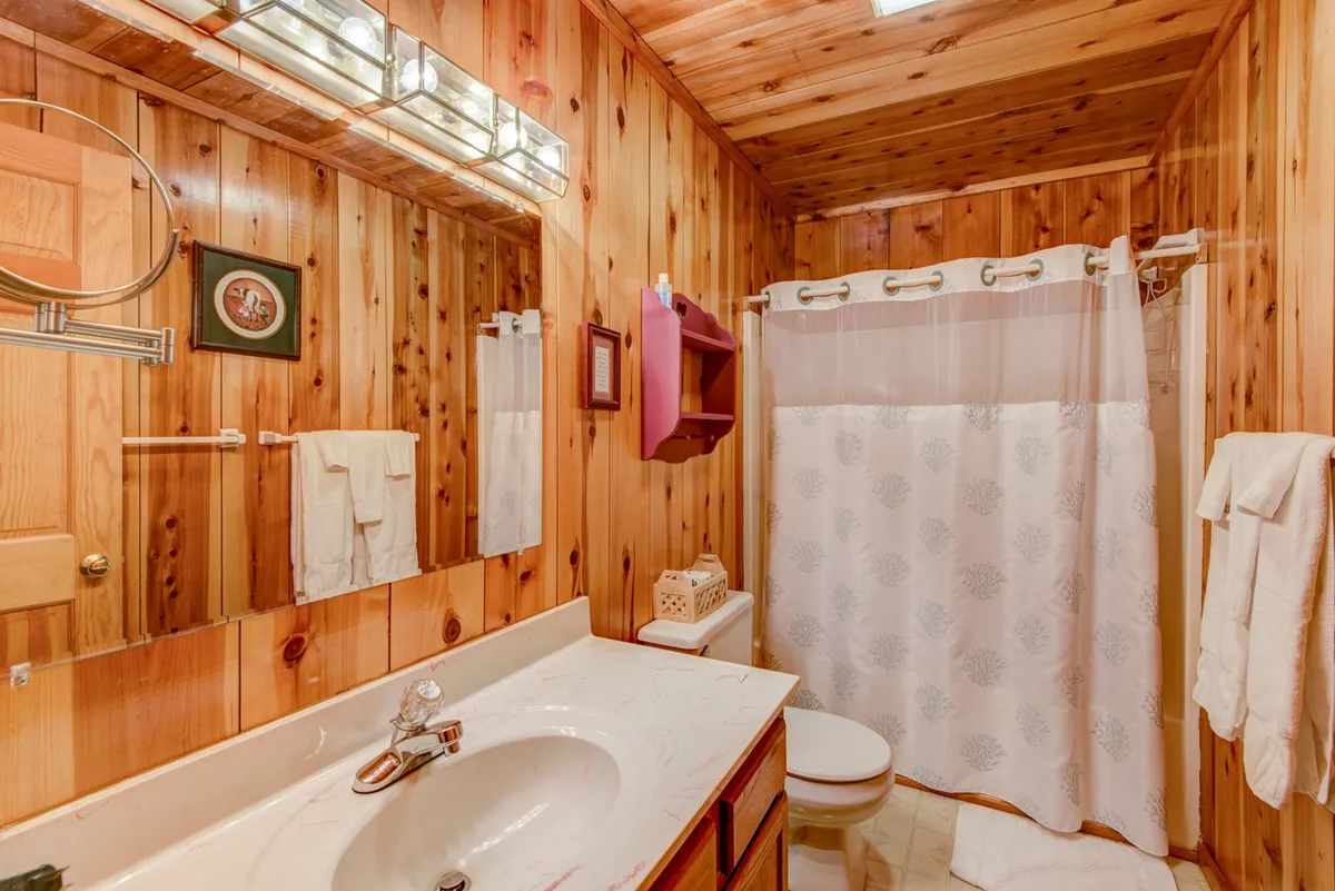 A rustic bathroom featuring wooden paneling, a wash basin, mirror, and a shower curtain. The inviting lighting and simple, practical layout make it clear this is a family-friendly, functional space.