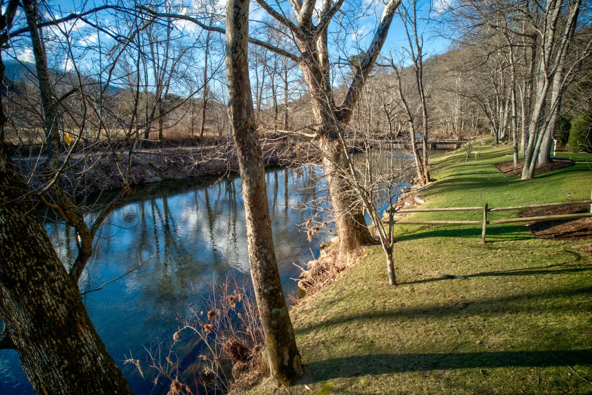 A tranquil river scene with gently flowing water bordered by lush grass and mature trees. The riverbank creates an inviting space perfect for picnicking, fishing, and nature walks, while the water's surface reflects the surrounding greenery and sky.