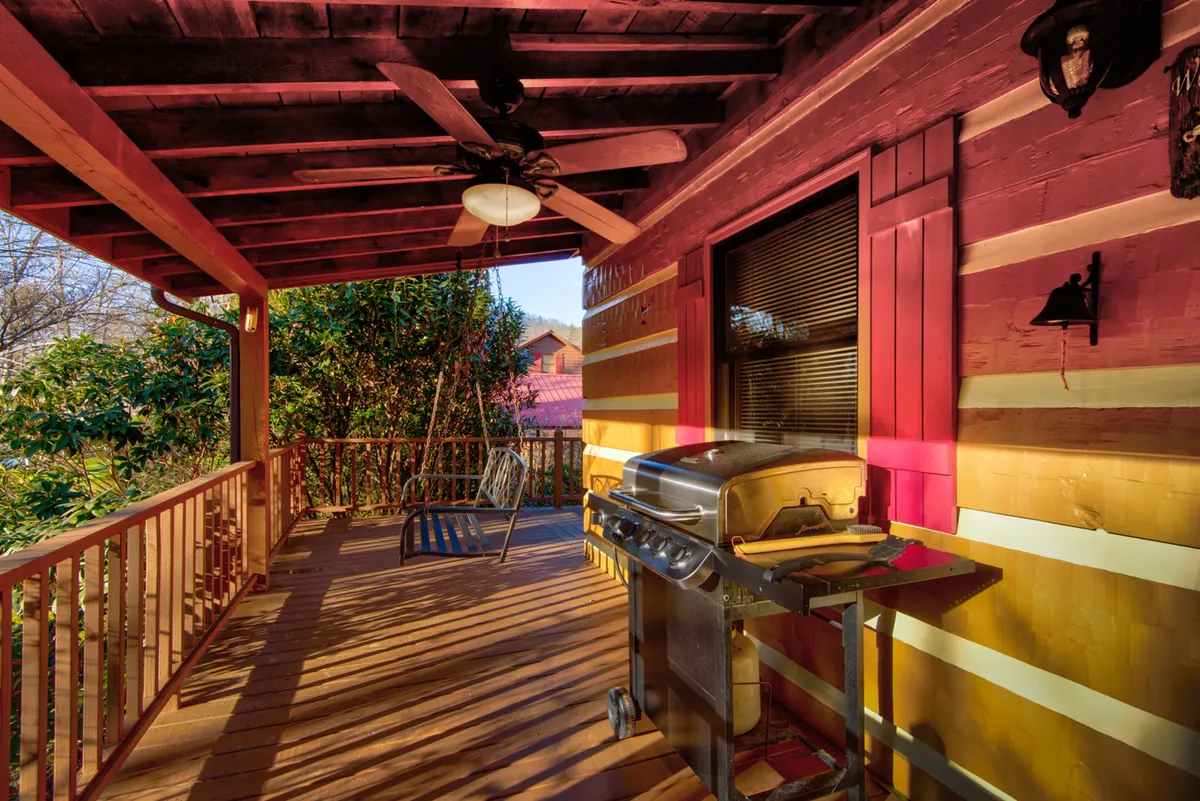 An exterior porch area with a gas grill, a comfortable swing chair, and sturdy wooden railings. The porch is bathed in soft light, suggesting late afternoon or early evening, and looks perfect for barbecuing, lounging, or enjoying mountain breezes.