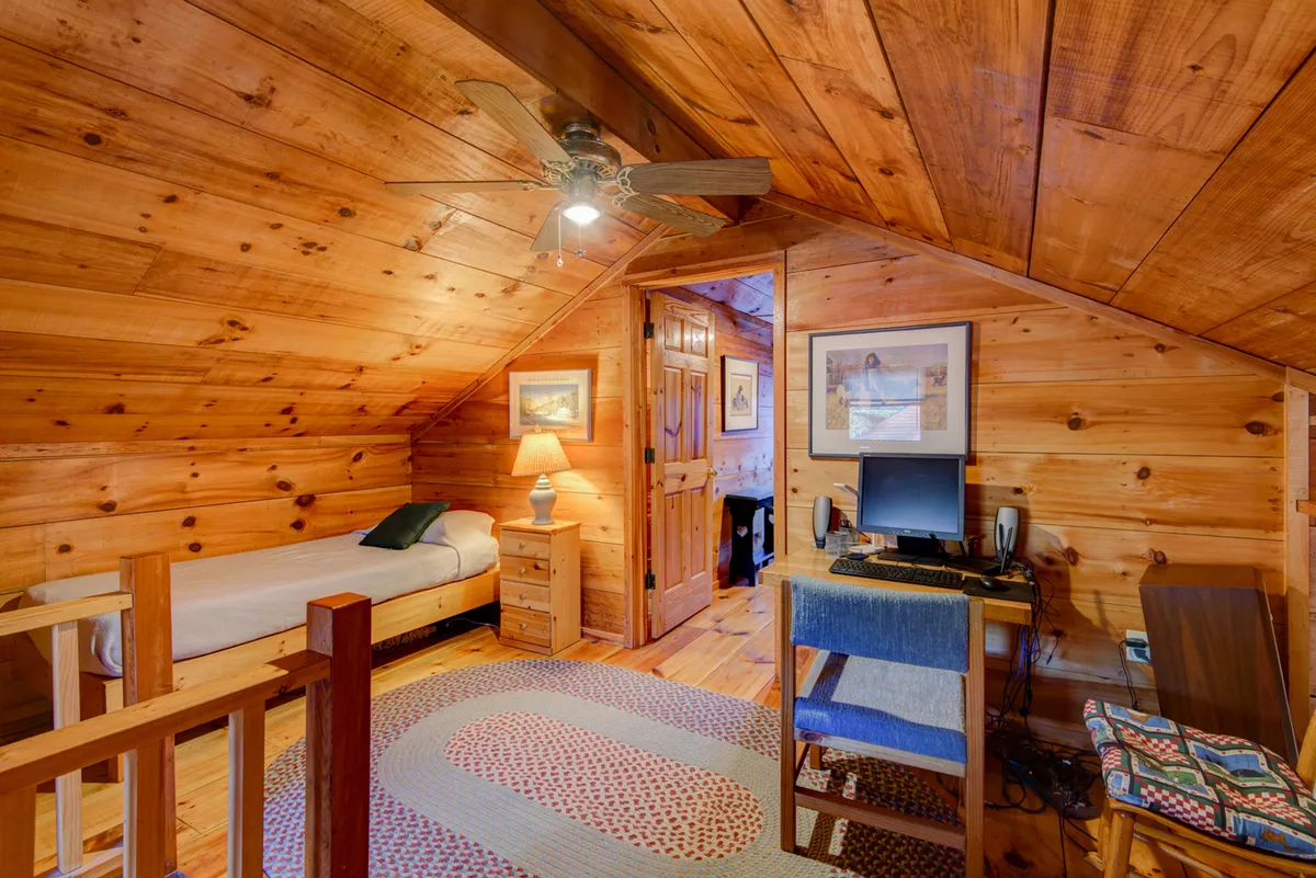 A warm and practical bedroom or study nook with a wooden ceiling, single bed, desk with computer, lamp, and rug. This cozy space offers a glimpse of everyday comforts, suggesting WiFi access, a place to work or relax, and family-friendly amenities.