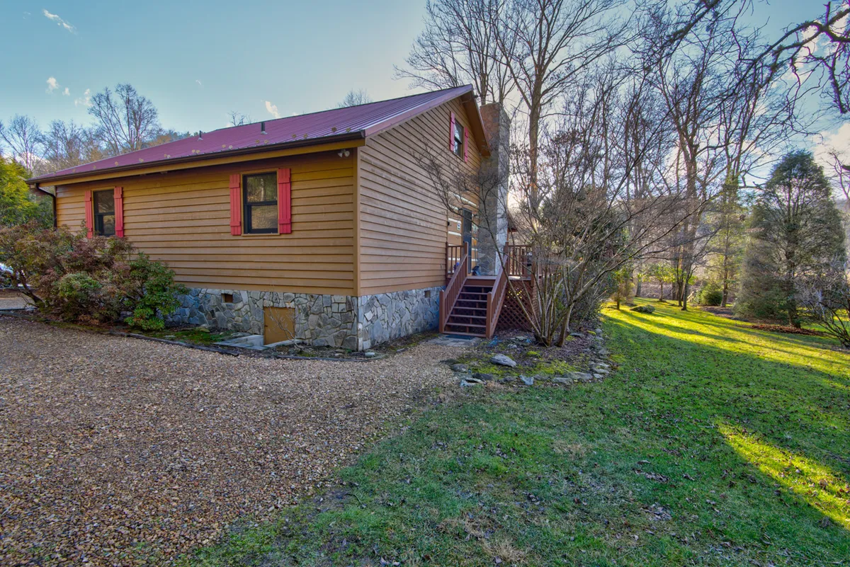 A warm and inviting living room with wooden siding and red shutters, featuring a gravel driveway and stone foundation surrounded by trees. The space exudes a homey, welcoming atmosphere, suggestive of a cabin nestled in nature. The large windows hint at possible river or nature views from within.