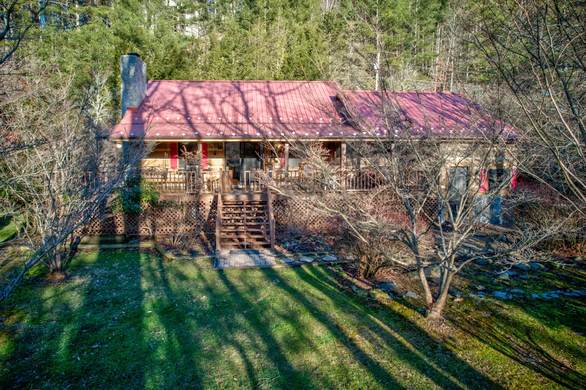 A charming view of the property's exterior, with a red roof, welcoming front porch with seating, and a stone pathway leading to the entrance. The home is surrounded by greenery and natural mountain scenery, creating an inviting first impression that speaks to the area's warmth and hospitality.