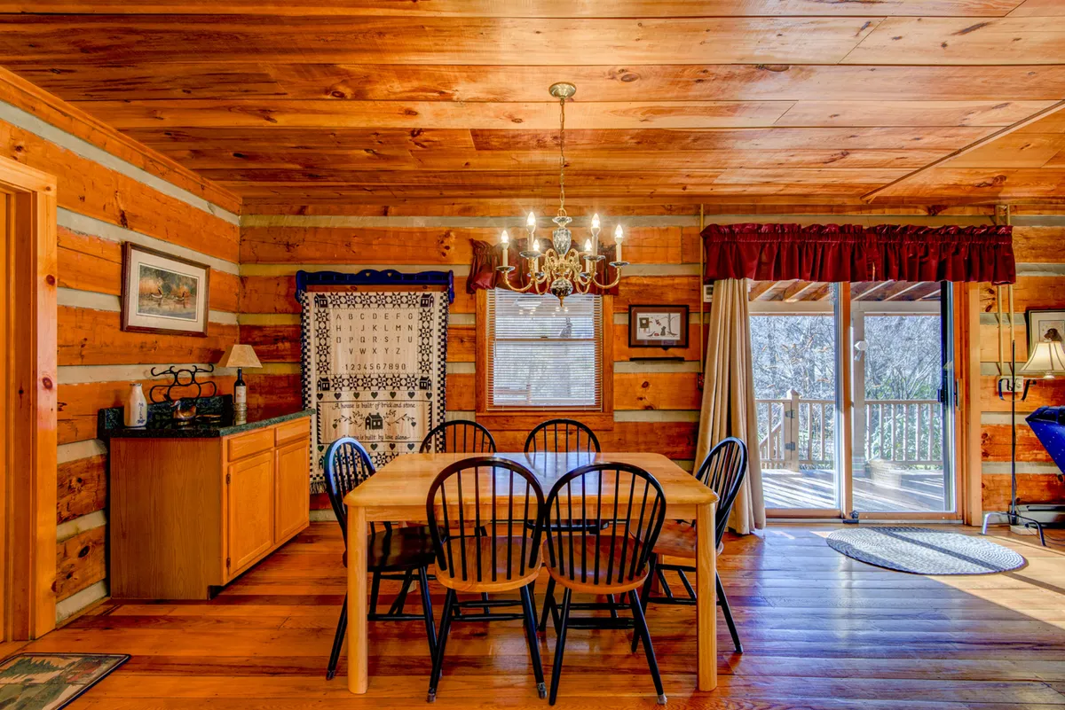 A dining area featuring a wooden table, elegant chandelier, wood-paneled walls, and a large sliding door that likely opens to views outside. The space feels intimate yet open—ideal for family meals or game nights, with natural light enhancing the welcoming ambiance.