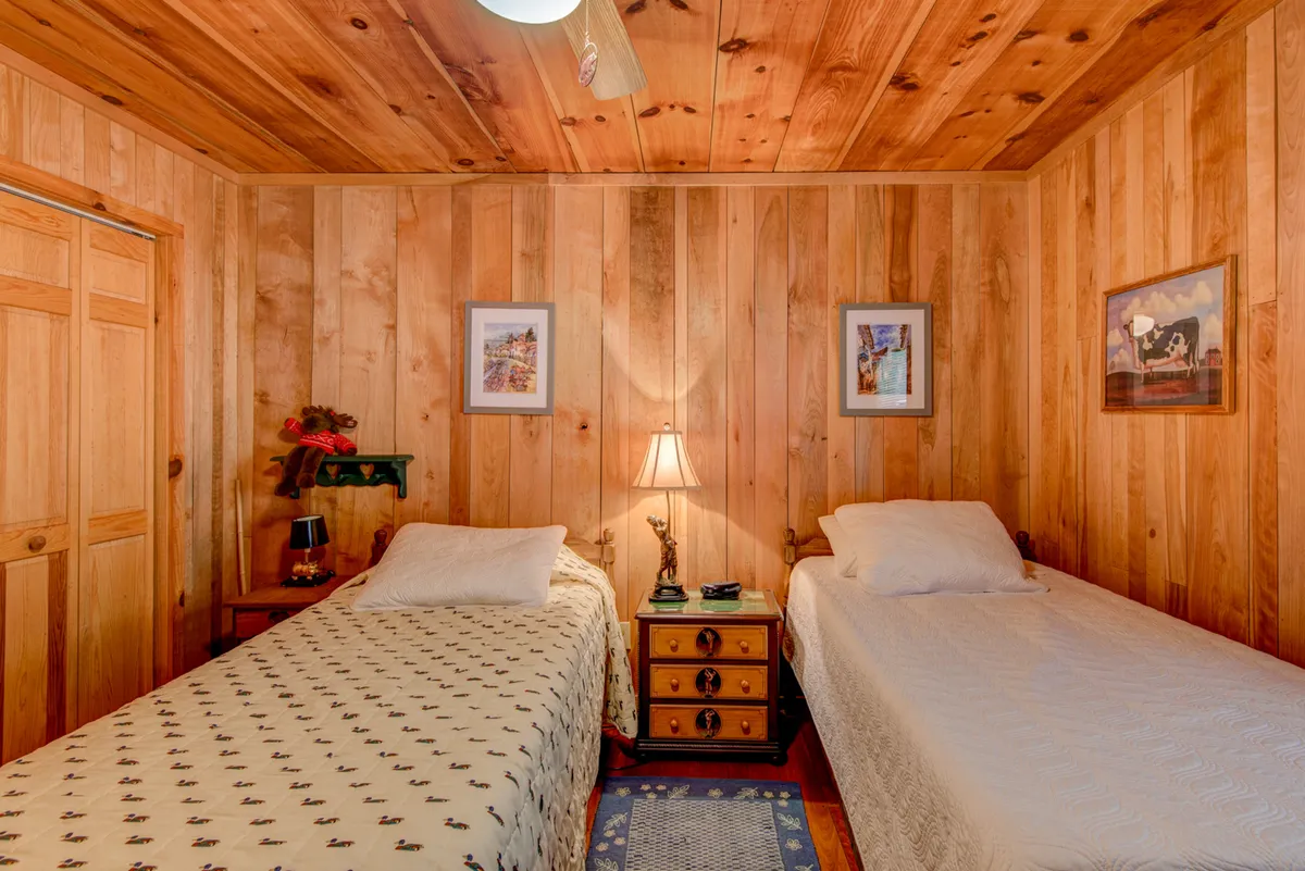 A warm, inviting bedroom with two beds, each dressed in plush linens. The wooden walls and soft lamp light create a cozy, restful atmosphere. Framed pictures add a personal touch, and the room feels like a perfect retreat for a peaceful night's sleep.
