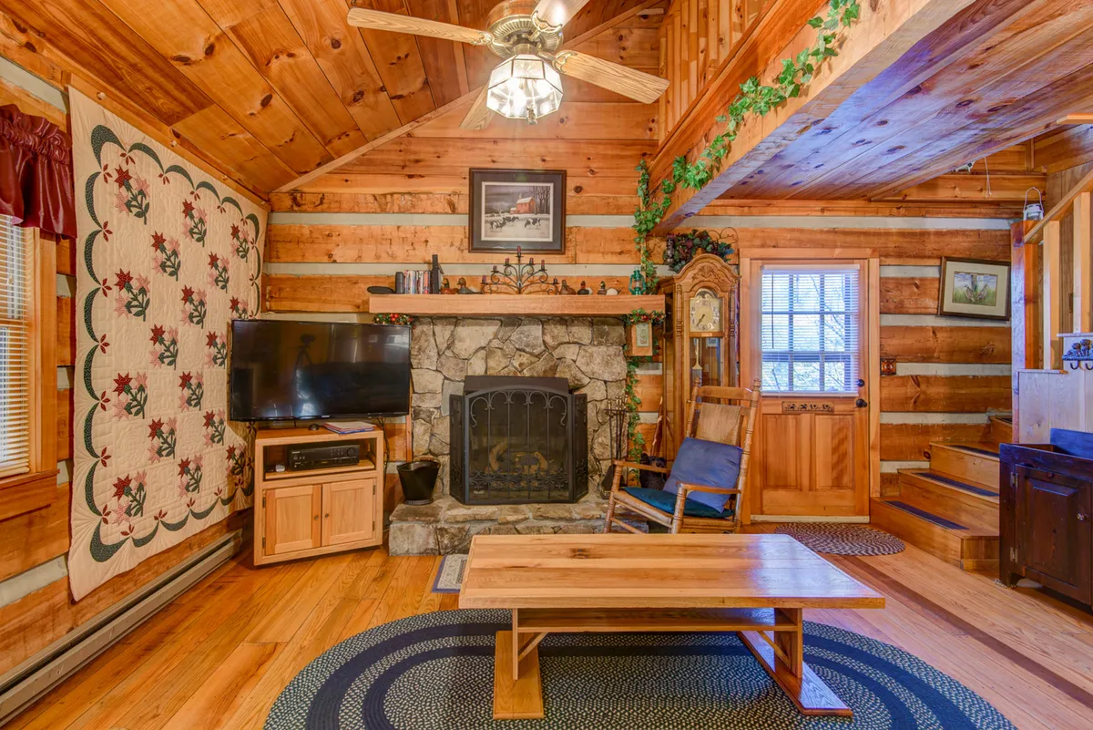A cozy living room anchored by a large stone fireplace, with inviting wooden furniture, a quilted wall hanging, and a TV. The space exudes warmth and comfort, perfect for relaxing after a day in the mountains, and hints at the home's reliable heating—a key amenity for mountain living.