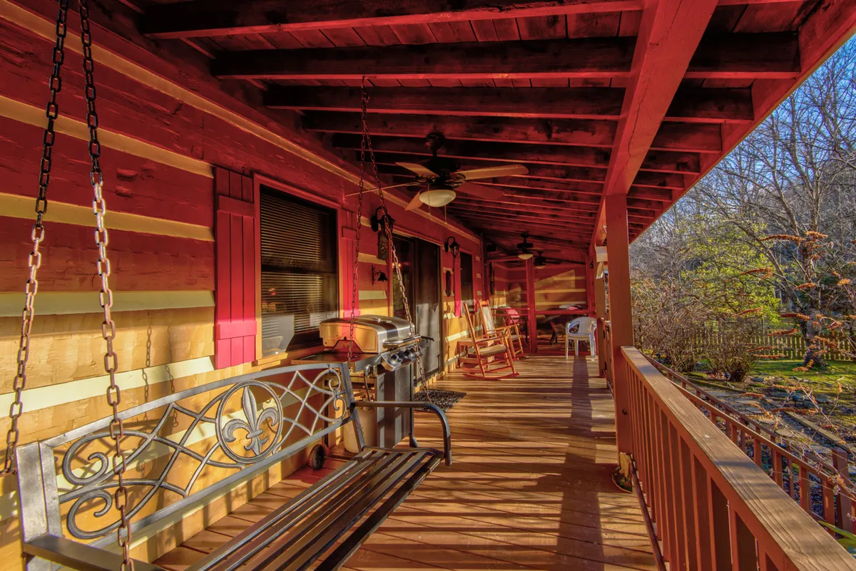 A welcoming outdoor porch featuring a swing bench, several rocking chairs, a gas grill, and warm wooden flooring, all surrounded by lush greenery. The scene radiates comfort and invites residents to enjoy nature while relaxing or socializing. The setting suggests both morning tranquility and convivial evening gatherings.