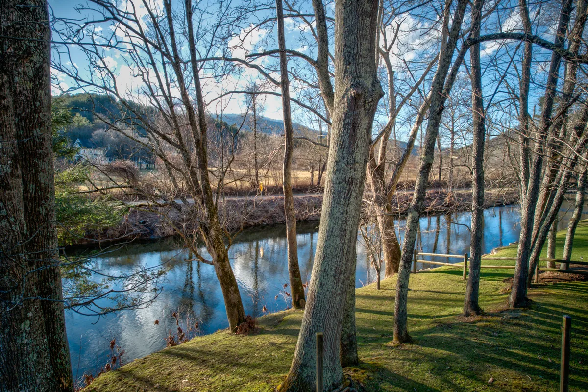 A serene riverside view with a calm river winding past tall trees and gentle hills in the background. The natural setting feels peaceful and secluded, perfect for fishing, birdwatching, or simply soaking in the scenery. The riverbank is accessible for outdoor activities and quiet moments.