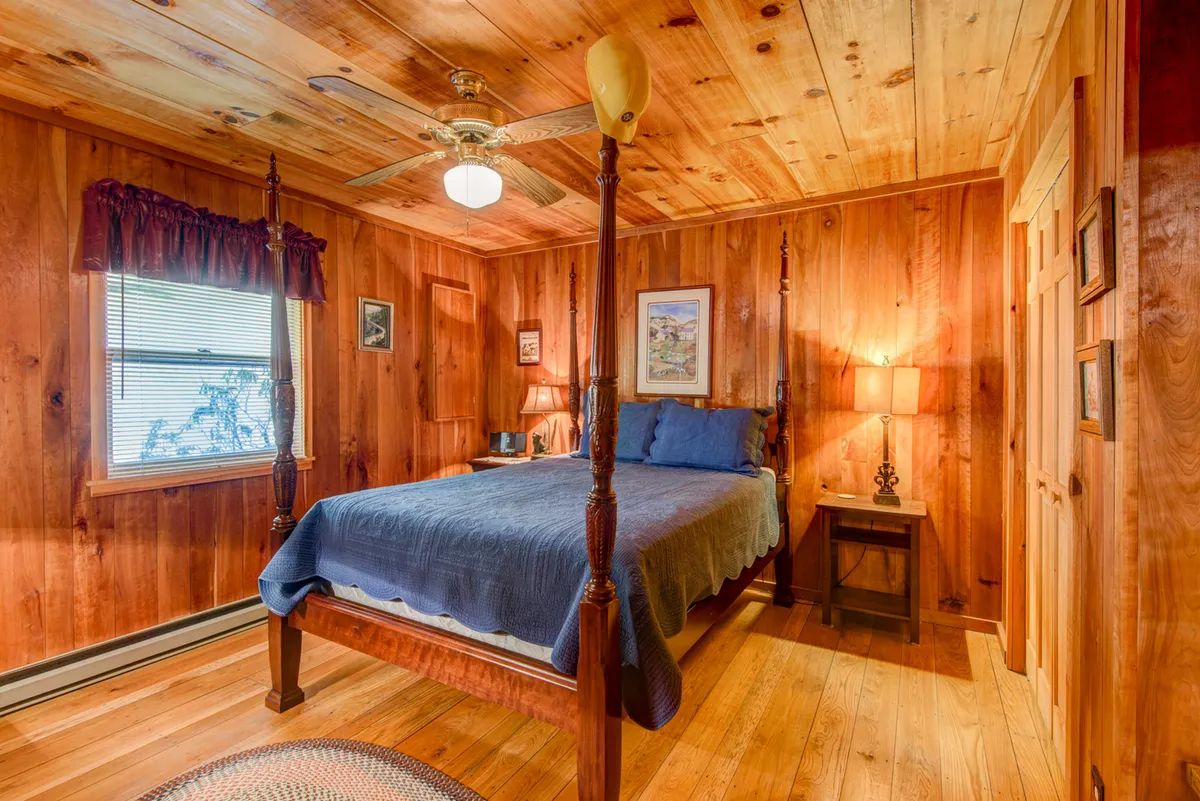 A charming bedroom featuring a cozy four-poster bed, wooden paneling, and a window with soft curtains. The nightstand, lamp, and gentle morning light create a serene, homey feel, underscoring the intimate retreat aspect.